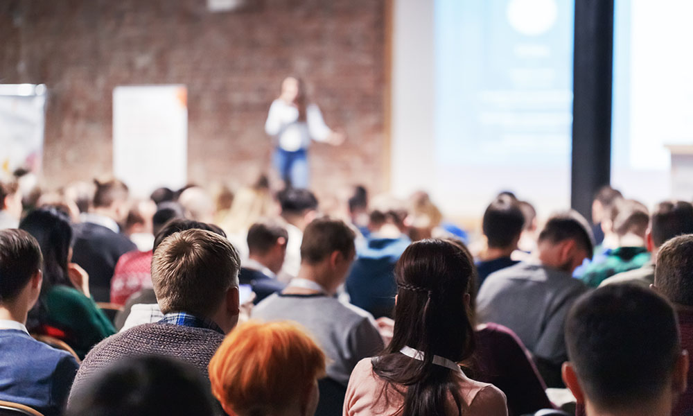 Upcoming Spring Event - View of a Large Group of People Attending a Conference Event with Views of a Female Speaker Presenting in the Front